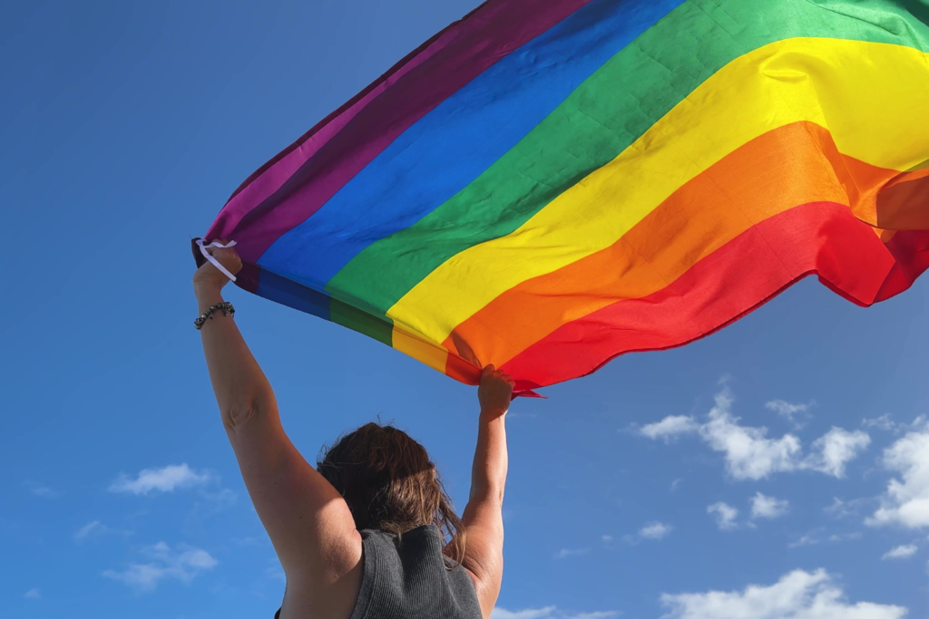 Person raises a large rainbow Pride flag against a clear blue sky, celebrating LGBTQ+ pride.
